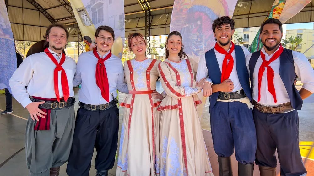 Grupo de dançarinos adultos em trajes gaúchos posa diante do painel do projeto
