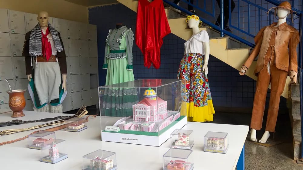 Manequins com vestidos e trajes regionais expostos junto a maquetes de arquitetura brasileira