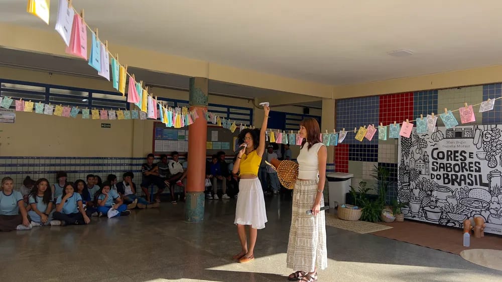 Apresentadoras ao lado do painel do projeto, no pátio decorado com varal de bandeirinhas