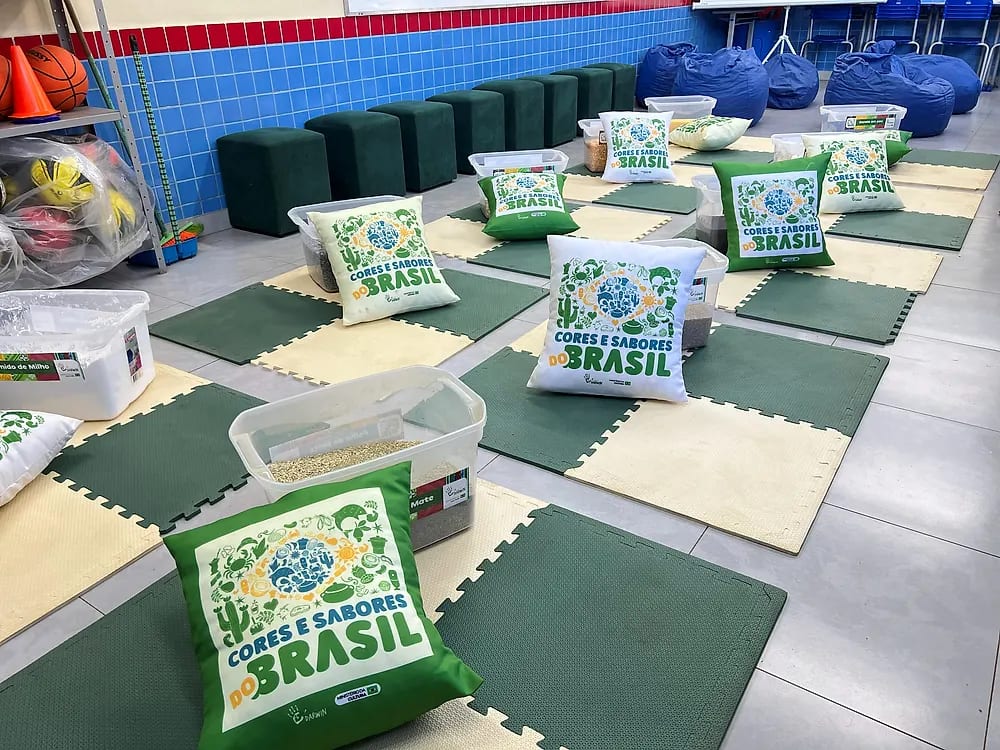 Circuito sensorial montado no chão: tapetes verdes e creme, almofadas com a marca do projeto e caixas transparentes com grãos para as crianças explorarem