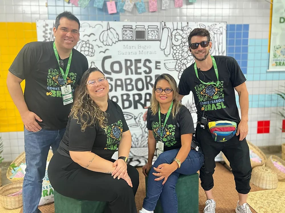 Integrantes da equipe do projeto, de camiseta preta com a marca Cores e Sabores do Brasil, posam sorrindo diante do painel ilustrado