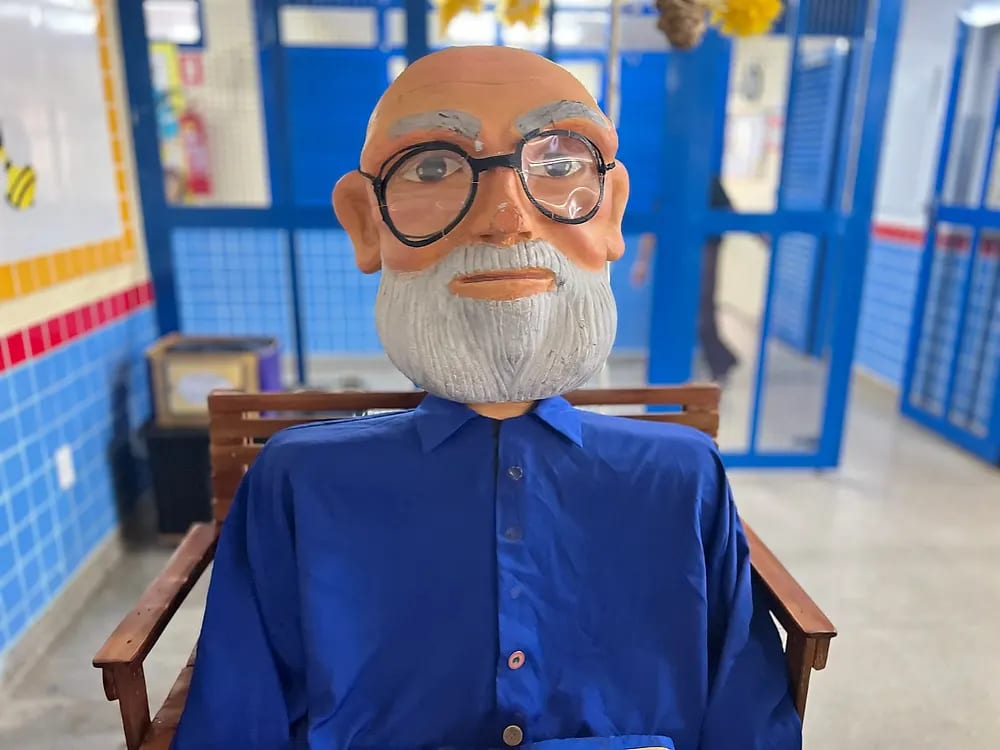 Boneco gigante de um senhor de óculos, barba branca e camisa azul, sentado em um banco de madeira no corredor da escola
